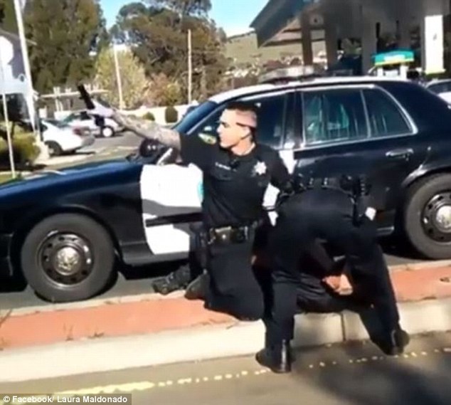 As witnesses begin to chant police brutality at the officer, he pulls out his gun on the crowd shouting at them to 'get the f*** back' (above)